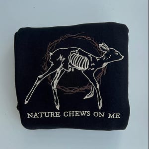 Puede incluir: Sudadera negra con una imagen bordada en blanco de un esqueleto de ciervo dentro de una corona. El texto "NATURE CHEWS ON ME" está bordado debajo de la imagen.