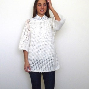 May include: A white crochet top with short sleeves and a loose, open weave design. The top is worn over a collared shirt with a light blue floral pattern. The person is wearing dark blue jeans.