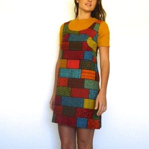Puede incluir: Un vestido de patchwork colorido con un patrón geométrico en tonos de rojo, azul, verde y marrón. El vestido tiene un diseño sin mangas y una silueta ajustada.