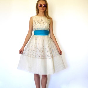 Puede incluir: Un vestido blanco sin mangas con estampado floral y una faja azul. El vestido tiene una falda completa y llega a la rodilla.