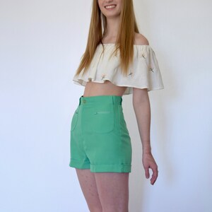 May include: A woman wearing a white off-the-shoulder top with floral embroidery and high-waisted green shorts.