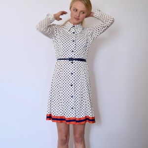 Puede incluir: Un vestido blanco con estampado de lunares azules y un cinturón negro. El vestido tiene cuello y mangas largas. El dobladillo del vestido tiene un ribete rojo y azul.