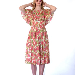 Può includere: Una donna indossa un abito rosa e verde a fiori con scollatura a barca e un colletto arricciato. Indossa occhiali da sole e ha le mani sulla testa.