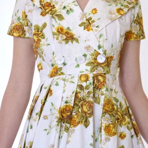 May include: A white dress with a yellow floral print and a double-breasted button closure. The dress has a collared neckline and short sleeves.
