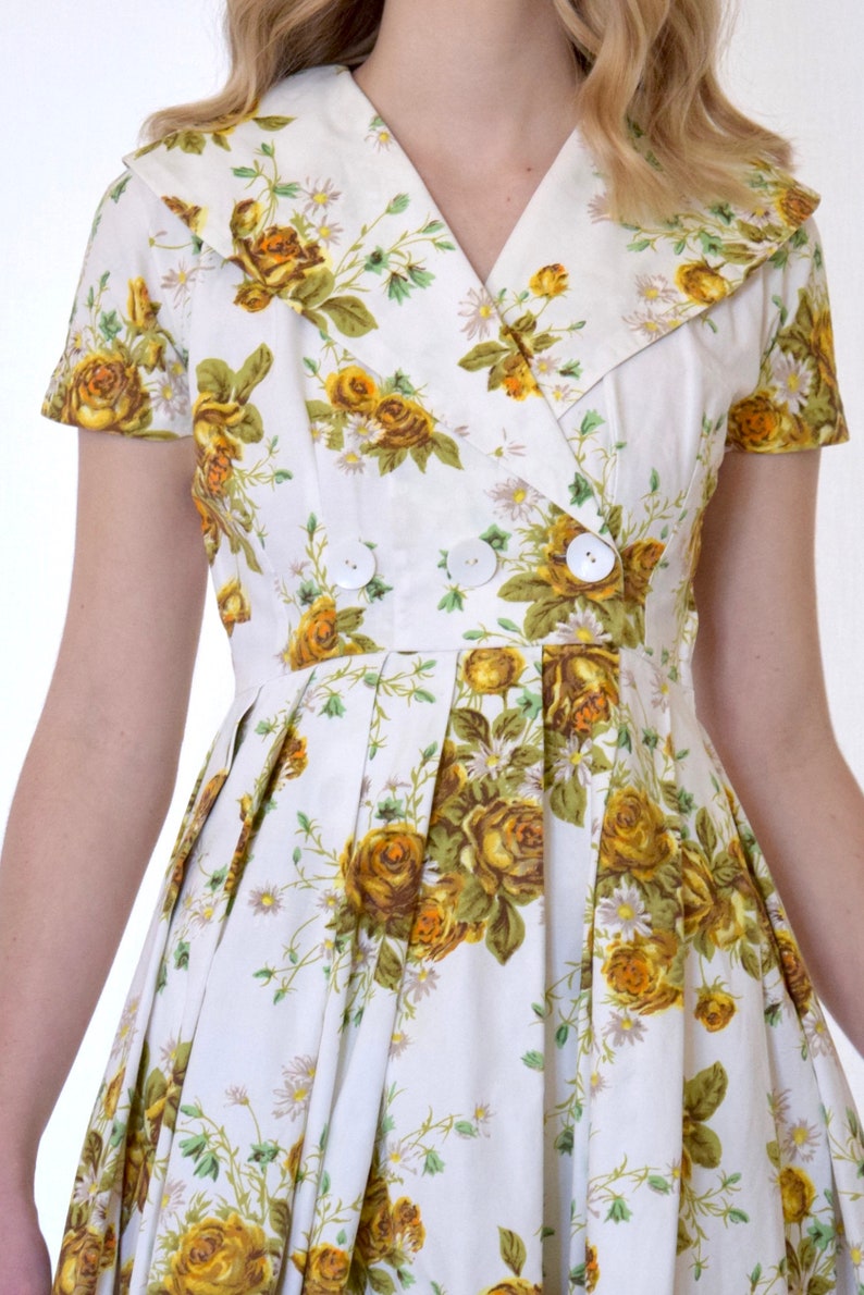 May include: A white dress with a yellow floral print and a double-breasted button closure. The dress has a collared neckline and short sleeves.