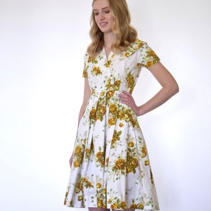 May include: A white dress with a yellow floral print and a collared neckline. The dress has short sleeves and a pleated skirt.