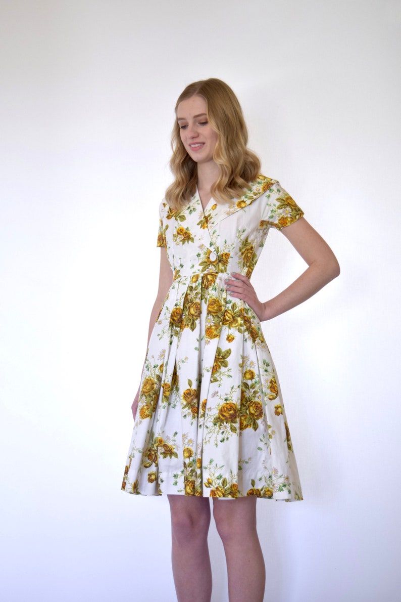 May include: A white dress with a yellow floral print and a collared neckline. The dress has short sleeves and a pleated skirt.