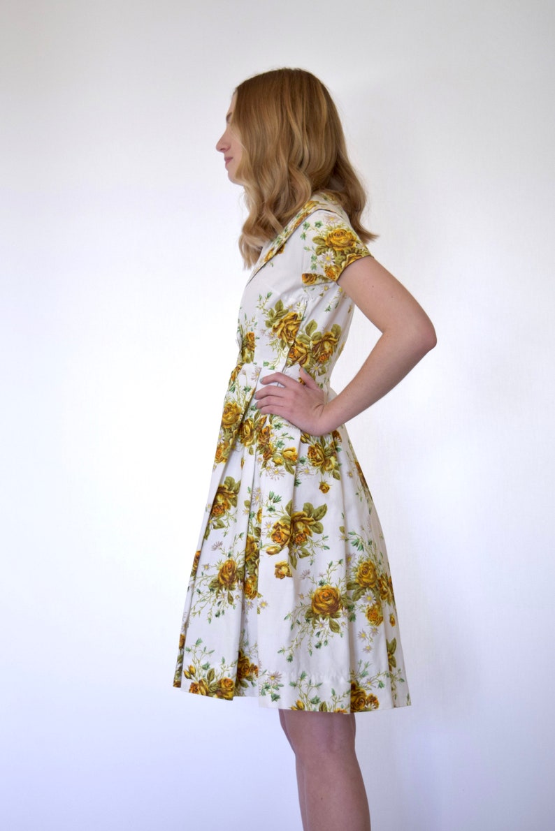 May include: A white dress with a yellow floral print. The dress has a collared neckline, short sleeves, and a pleated skirt.