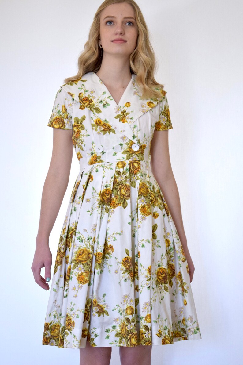 May include: A white dress with a yellow floral print and a large collar. The dress has a button closure and a pleated skirt.