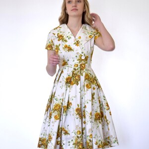 May include: A white dress with a yellow floral print and a collared neckline. The dress has short sleeves and a pleated skirt.