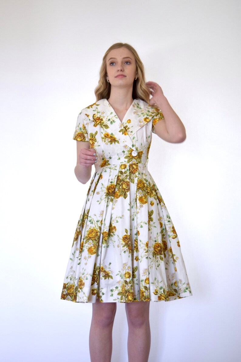 May include: A white dress with a yellow floral print and a collared neckline. The dress has short sleeves and a pleated skirt.