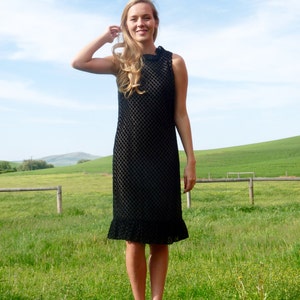 Könnte beinhalten: Eine Frau trägt ein schwarzes, ärmelloses, knielanges Kleid mit einem strukturierten Muster. Sie steht auf einem Feld mit grünem Gras und einem Holzzaun im Hintergrund.