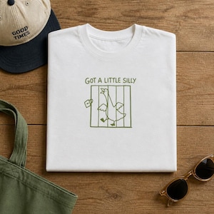 May include: A white t-shirt featuring a green line-art graphic of a goose inside a cage with an open door and a padlock. Above the cage, the text reads "GOT A LITTLE SILLY" in green. The design is centered on the chest of the folded garment.