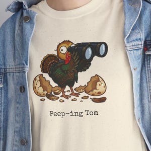 May include: A cream-colored t-shirt featuring a graphic of a brown turkey emerging from a cracked egg and looking through black binoculars. Below the turkey, the text reads Peep-ing Tom. The shirt is worn under a blue denim jacket.