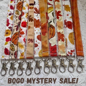 May include: A collection of ten fabric lanyards featuring autumn-themed patterns with pumpkins, red and orange leaves, and berries. Each strap is finished with a silver-toned metal swivel lobster clasp at the bottom for secure attachment.