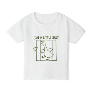 May include: A white short-sleeved t-shirt featuring a dark green line-art graphic of a goose inside a cage with an open door and a padlock. Above the cage, the text reads "GOT A LITTLE SILLY" in a simple, dark green, hand-drawn style font.