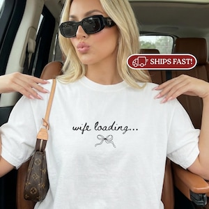 May include: A white short-sleeved t-shirt featuring the black text "wife loading..." centered on the chest, accompanied by a simple black line drawing of a bow underneath the text. The shirt has a classic crew neckline and a relaxed fit.