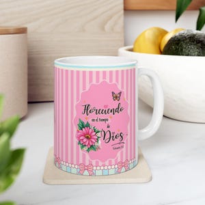 Könnte beinhalten: Ein weißer Keramikbecher mit einem rosa-weiß gestreiften Muster und einem gewellten Etikett in der Mitte. Das Etikett zeigt eine rosa Blume, einen Schmetterling und den Text Floreciendo en el tiempo de Dios Eclesiastés 3:11.