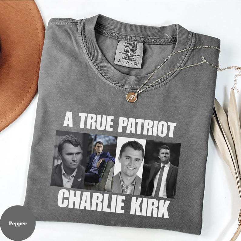 K&ouml;nnte beinhalten: Ein graues Comfort Colors T-Shirt mit dem Text "A True Patriot Charlie Kirk" und vier Bildern von Charlie Kirk. Eine goldene Halskette und ein brauner Hut sind ebenfalls sichtbar.