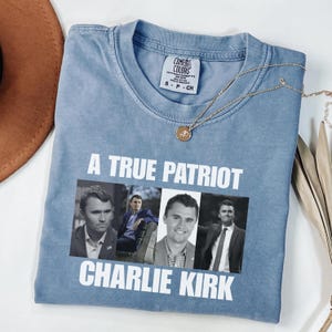 K&ouml;nnte beinhalten: Hellblaues Comfort Colors T-Shirt mit dem wei&szlig;en Text "A TRUE PATRIOT CHARLIE KIRK". Das Shirt zeigt vier Bilder von Charlie Kirk. Eine goldene Halskette und ein brauner Hut sind ebenfalls zu sehen.