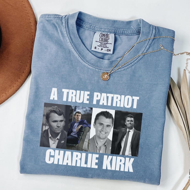 K&ouml;nnte beinhalten: Hellblaues Comfort Colors T-Shirt mit dem wei&szlig;en Text "A TRUE PATRIOT CHARLIE KIRK". Das Shirt zeigt vier Bilder von Charlie Kirk. Eine goldene Halskette und ein brauner Hut sind ebenfalls zu sehen.
