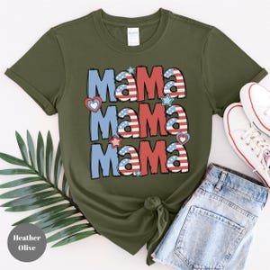 May include: Heather olive green t-shirt with the word "Mama" repeated three times in a patriotic design. The word "Mama" is in red, white, and blue colors with heart and star accents. The shirt is paired with denim shorts and white sneakers.
