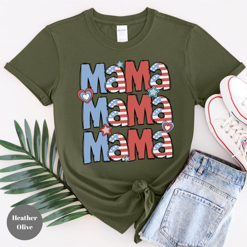 May include: Heather olive green t-shirt with the word "Mama" repeated three times in a patriotic design. The word "Mama" is in red, white, and blue colors with heart and star accents. The shirt is paired with denim shorts and white sneakers.