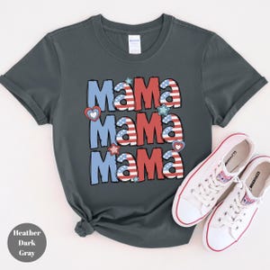 May include: Heather dark gray t-shirt with the word "Mama" repeated three times in red, white, and blue, with heart and star accents. White sneakers with red trim are next to the shirt. The shirt is made of a soft material.