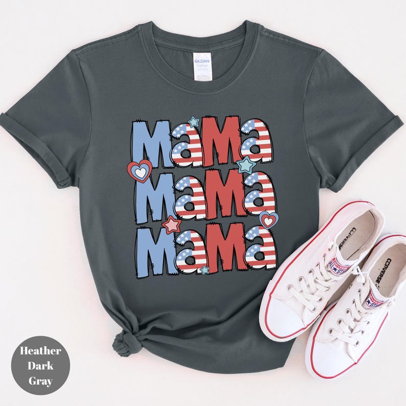 May include: Heather dark gray t-shirt with the word "Mama" repeated three times in red, white, and blue, with heart and star accents. White sneakers with red trim are next to the shirt. The shirt is made of a soft material.