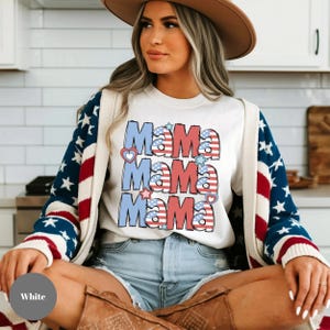 May include: White t-shirt with the word "Mama" repeated three times in red, white, and blue, with heart and star accents. The shirt is paired with denim shorts and a cardigan with a star-spangled design.