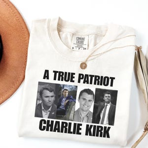 K&ouml;nnte beinhalten: Cremefarbenes T-Shirt mit dem Text "A True Patriot Charlie Kirk" und drei Bildern von Charlie Kirk. Eine goldene Halskette und ein brauner Hut sind ebenfalls sichtbar.