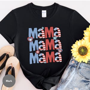 May include: Black t-shirt with the word "Mama" in red, white, and blue, with heart and star accents. The letters are designed to resemble the American flag. The shirt is paired with denim shorts and sandals.