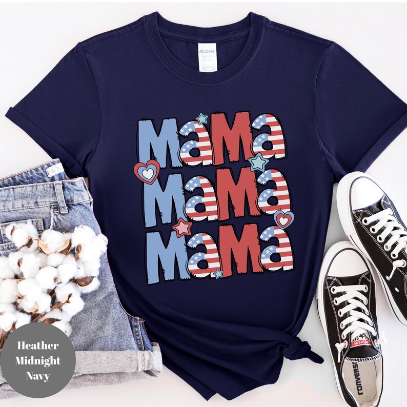May include: Navy blue t-shirt with the word "Mama" repeated three times in a patriotic design. The word "Mama" is in red, white, and blue colors with heart and star accents. The shirt is paired with denim shorts and black sneakers.