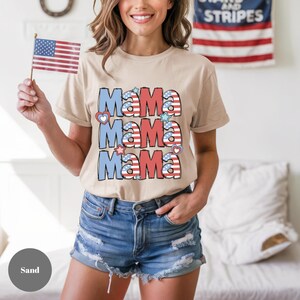 May include: Sand-colored t-shirt with the word "Mama" repeated three times in red, white, and blue, with heart and star accents. The shirt is paired with denim shorts. An American flag is held in the left hand.