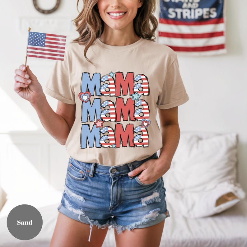 May include: Sand-colored t-shirt with the word "Mama" repeated three times in red, white, and blue, with heart and star accents. The shirt is paired with denim shorts. An American flag is held in the left hand.