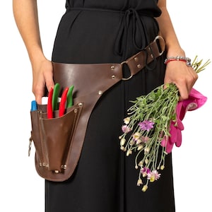 Puede incluir: Un cinturón de jardinería de cuero marrón con bolsillos que contienen herramientas rojas, verdes y azules. El cinturón se lleva sobre un vestido negro. Una mano sostiene un ramo de flores y un guante rosa.