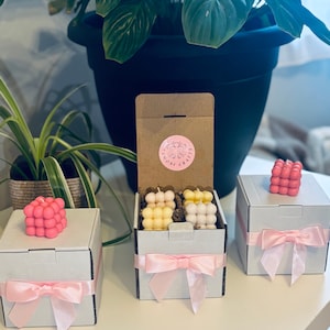 May include: Three white gift boxes with pink ribbons. The boxes contain small, colourful, cube-shaped candles. The box in the centre is open and shows the candles inside. The text on the box reads "The Royal Crafts".
