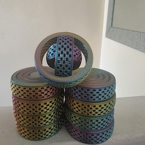 May include: Several iridescent, perforated, circular objects are stacked, with a matching sphere and ring. The objects have a metallic sheen, with colours shifting from blue to green to gold. The design features small, evenly spaced holes.