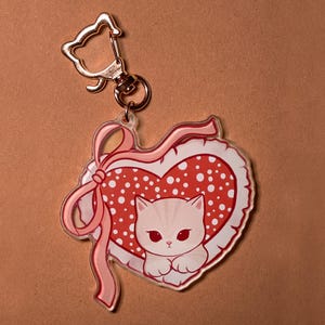 Op de afbeelding: Een heldere acryl sleutelhanger met een witte kat in een rood hart met witte stippen. Het hart is wit omlijnd en heeft een roze lint. De sleutelhanger heeft een goudkleurige sluiting.