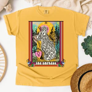 Może przedstawiać: Żółta koszulka z kolorową ilustracją karty tarota Cesarzowej. Karta przedstawia kobietę siedzącą na tronie, w koronie i trzymającą symbol płci żeńskiej. Tekst "THE EMPRESS" jest nadrukowany pod obrazkiem.