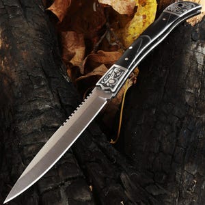 May include: A silver and black knife with a serrated edge and decorative silver accents. The handle is black with silver rivets. The knife is set against a dark, textured background with autumn leaves.