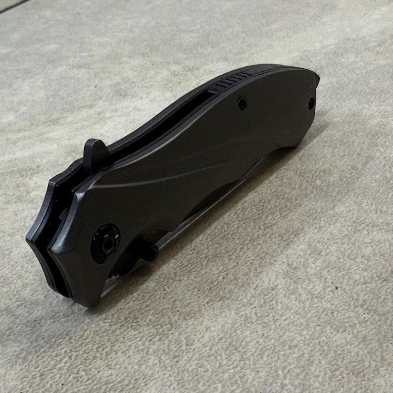 May include: A closed, dark gray folding knife with a curved handle and a textured back. The knife has a small, protruding tab for opening and a black pivot screw. The blade is not visible in this image.