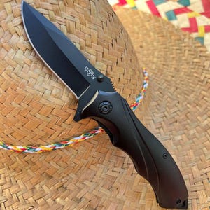 May include: A black folding knife with a sharp blade and a textured handle. The knife features a logo on the blade and is shown against a woven straw hat with a colorful trim.