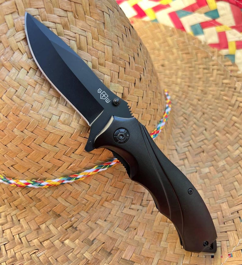 May include: A black folding knife with a sharp blade and a textured handle. The knife features a logo on the blade and is shown against a woven straw hat with a colorful trim.