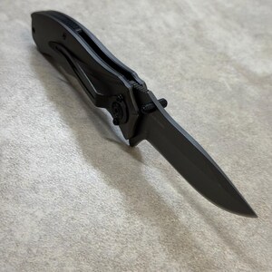May include: A matte black folding knife with a pointed blade. The knife features a clip for pocket carry and a locking mechanism. The handle and blade are a matching dark gray color.