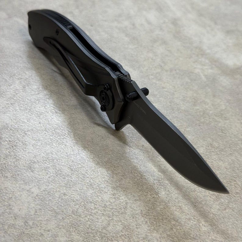 May include: A matte black folding knife with a pointed blade. The knife features a clip for pocket carry and a locking mechanism. The handle and blade are a matching dark gray color.