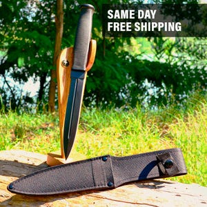 May include: A black tactical knife with a serrated edge, displayed in a wooden stand. The knife has a textured handle and is paired with a black sheath. The text "SAME DAY FREE SHIPPING" is visible in the background.
