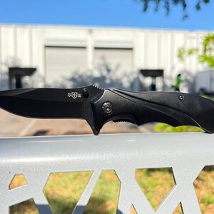 May include: A black folding knife with a sharp blade and a textured handle. The knife has a clip for easy carrying and the letters "G W" are visible on the blade. The knife is shown outdoors.