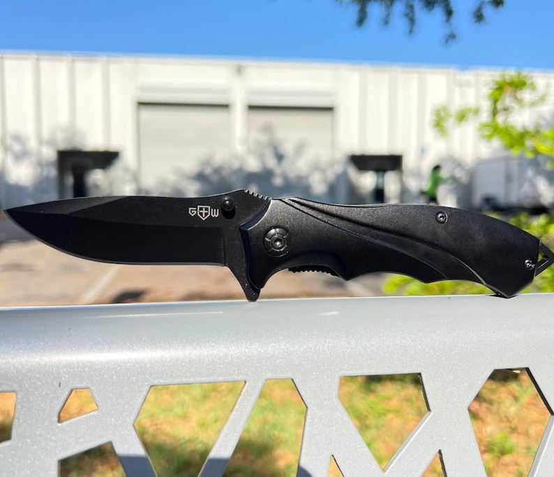 May include: A black folding knife with a sharp blade and a textured handle. The knife has a clip for easy carrying and the letters "G W" are visible on the blade. The knife is shown outdoors.