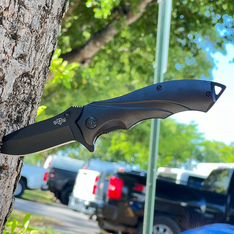 May include: A black folding knife with a partially serrated blade. The knife features a textured handle and a blade that is approximately 3.5 inches long. The knife is shown against a natural background.